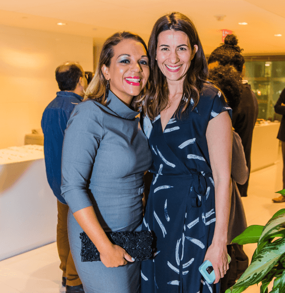 Two women standing together indoors at an event, smiling at the camera. Other people are visible in the background.