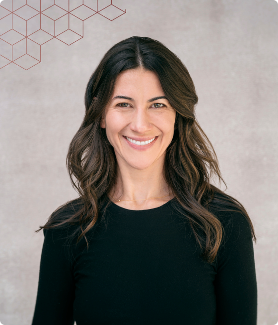 Woman with long brown hair wearing a black top, smiling in front of a light gray background with a geometric pattern in the top left corner.