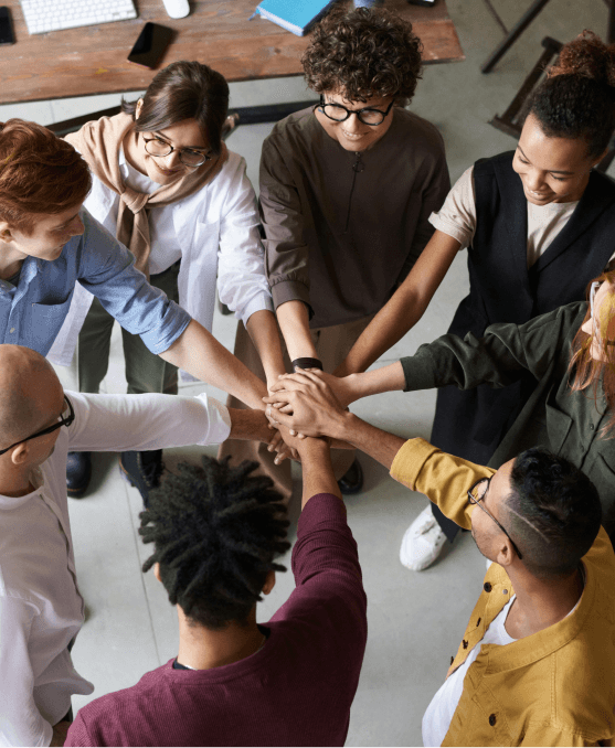 A group of eight people standing in a circle join hands in the center, showing teamwork and unity in a casual indoor setting.
